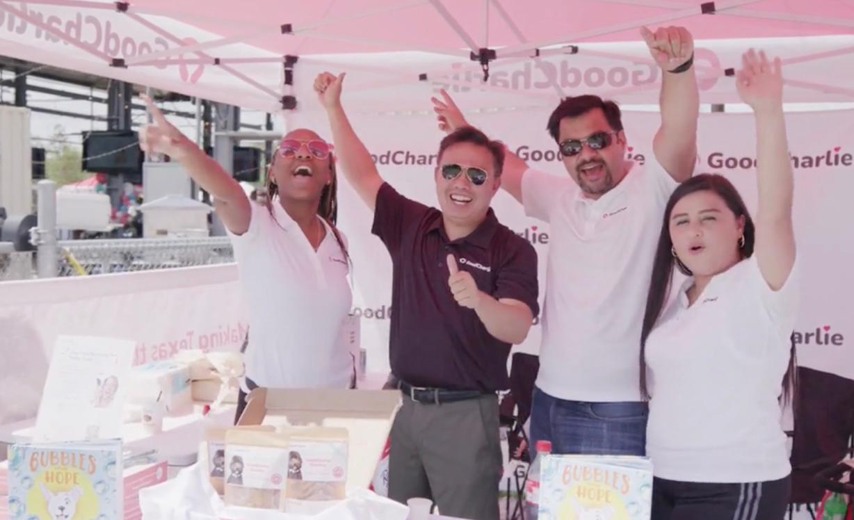 Four GoodCharlie team members are at an outdoor event, they are standing under a GoodCharlie branded gazebo cheering with their hands raised up.