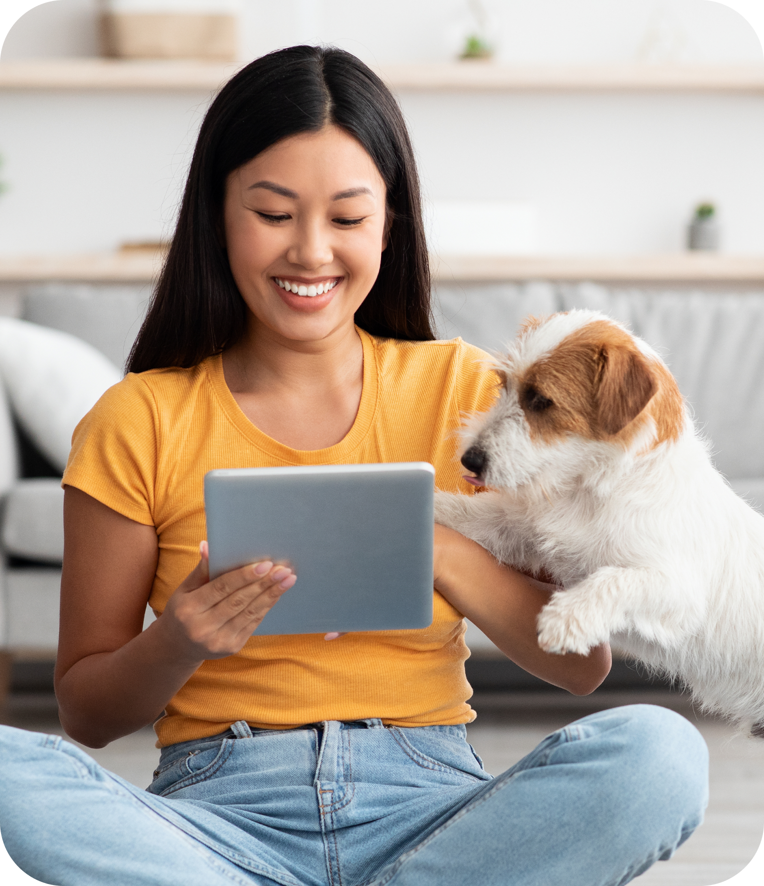 A woman smiling while sitting on the floor and pressing a tablet device in her hand. A dog is beside her peering into the tablet screen.