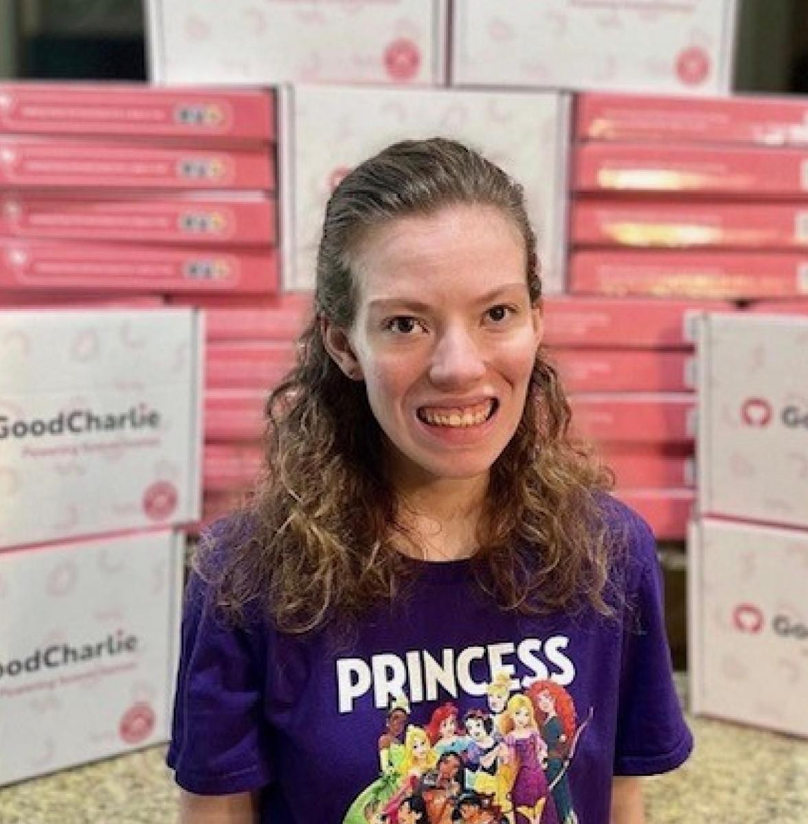 Jess is smiling, wearing a purple t-shirt with 'PRINCESS' inscribed in the front with a picture of Disney princesses. There are boxes of GoodCharlie treats in the background.